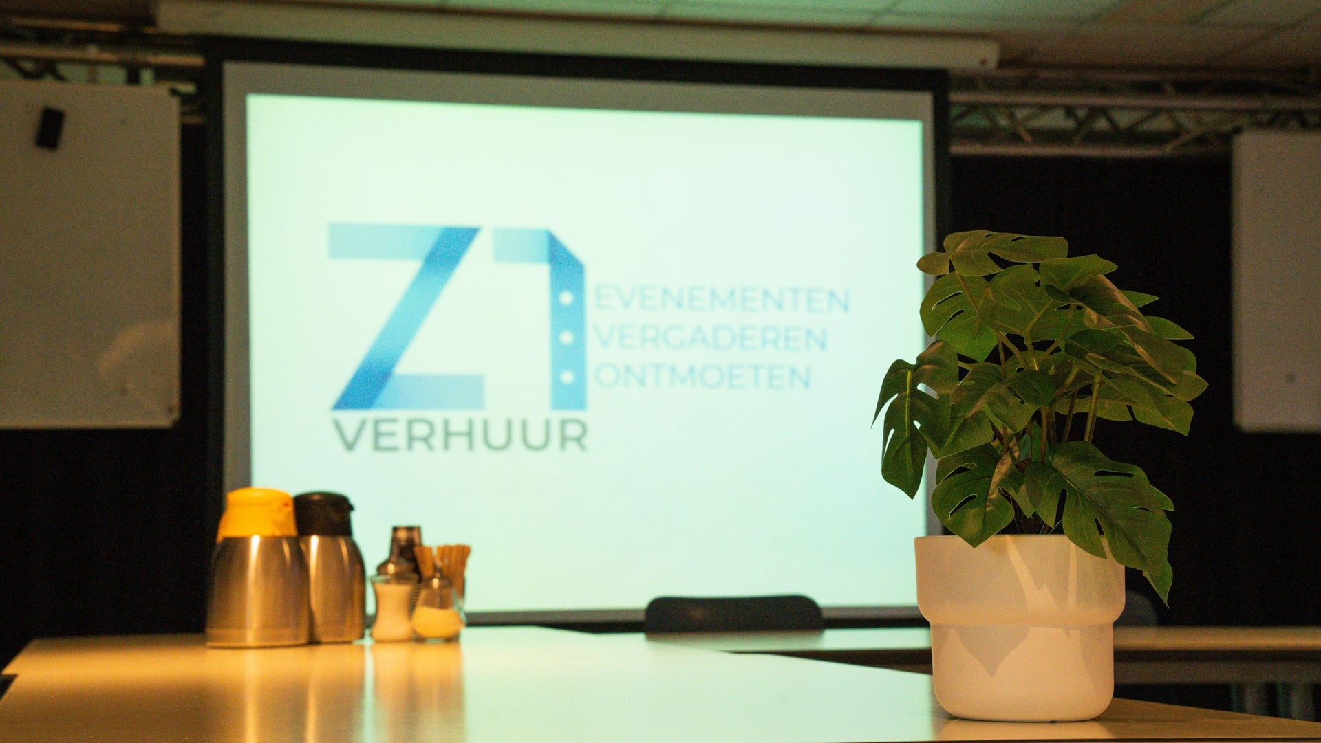 Detail van de vergadertafel: koffiekannen, roerstaafjes en een plant voor het projectiescherm met het logo van Z1 Verhuur