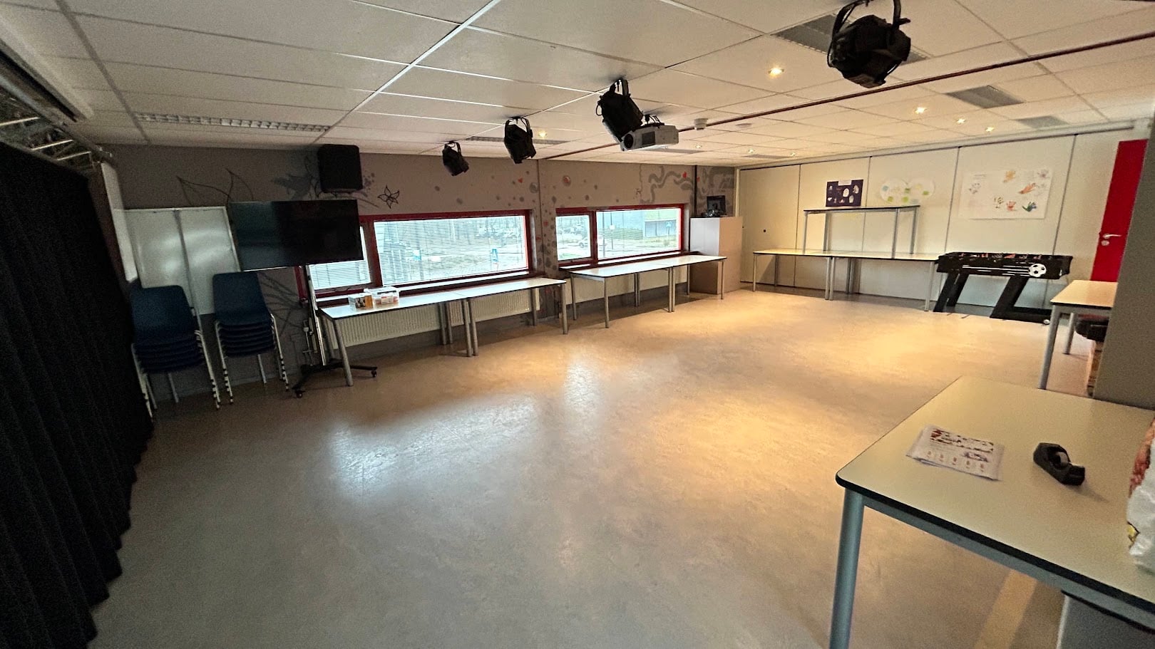 Dezelfde ruimte leeg: vrije vloer, verrijdbare tafels langs de wand, tv-scherm, beamer en spots aan een truss